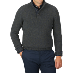 A person wearing a Gran Sasso Green Melange Wool Quarter Button Sweater paired with dark blue trousers is standing with one hand in a trouser pocket against a grey background.