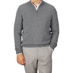 A person wearing a Gran Sasso Grey Melange Wool Cashmere 1/4 Zip Sweater over a white shirt and light grey pants stands with one hand in their pocket. The solid gray background complements the sustainable knitwear, creating a cohesive look.