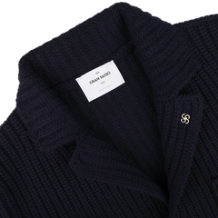 Close-up of a navy blue chunky knitted wool cardigan by Gran Sasso, featuring a metallic pin on the lapel and a white label inside bearing the brand name.