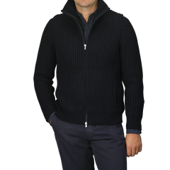 A person wearing a navy blue heavy knit cardigan made of wool and cashmere by Gran Sasso over a dark shirt and pants, with hands in pockets, stands against a plain gray background.