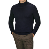 A man wearing a Navy Rain Wool Rib Roll Neck by Gran Sasso and brown pants stands with his hands in his pockets against a plain background.