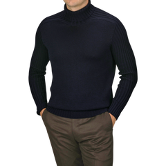 A man wearing a Navy Rain Wool Rib Roll Neck by Gran Sasso and brown pants stands with his hands in his pockets against a plain background.