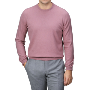 A person wearing a Pale Pink Cashmere Crewneck Sweater by Gran Sasso and gray pants stands with one hand in their pocket against a gray background.