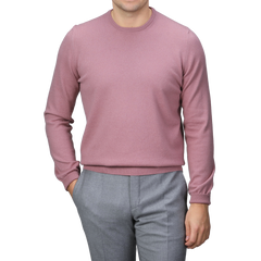 A person wearing a Pale Pink Cashmere Crewneck Sweater by Gran Sasso and gray pants stands with one hand in their pocket against a gray background.