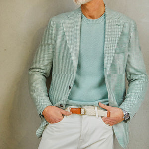 A person wearing a Baltzar Sartorial green houndstooth wool silk linen blazer, a pale turquoise sweater, white pants, and a wristwatch, with hands casually placed in the pockets.