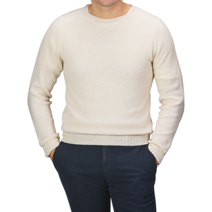 A person wearing the Almond White Extrafine Lambswool Crewneck by Harley of Scotland and dark pants stands with hands in their pockets against a gray background.