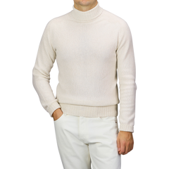 A person wearing a Harley of Scotland Almond White Extrafine Lambswool Rollneck and white pants stands against a neutral background.