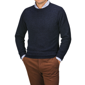 A person stands with hands in pockets, wearing a Harley of Scotland Cosmos Blue Extrafine Lambswool Crewneck over a light shirt and brown pants, set against a neutral background.