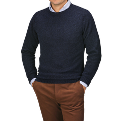 A person stands with hands in pockets, wearing a Harley of Scotland Cosmos Blue Extrafine Lambswool Crewneck over a light shirt and brown pants, set against a neutral background.