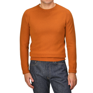 A person wearing the Sienna Orange Extrafine Lambswool Crewneck by Harley of Scotland and blue jeans stands against a gray background.