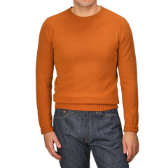 A person wearing the Sienna Orange Extrafine Lambswool Crewneck by Harley of Scotland and blue jeans stands against a gray background.