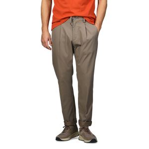 Dressed in an orange shirt, a person stands with hands in pockets, wearing sneakers and dark beige nylon stretch drawstring trousers by Herno.