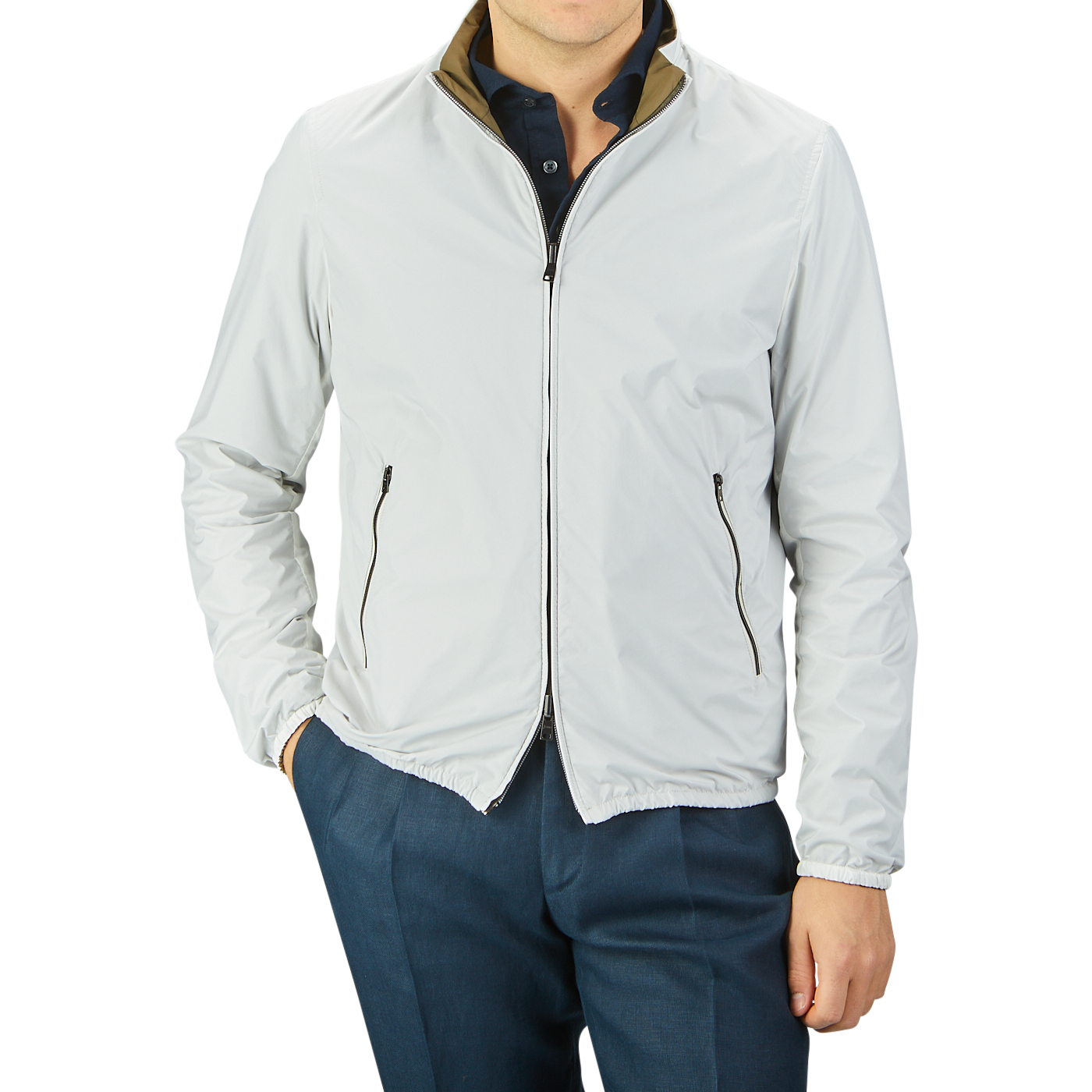 A person wearing a light gray, slim-fit zip-up jacket and dark blue pants is standing with hands in pockets. The Herno Olive Green White Reversible Nylon Blouson, made of water-resistant nylon, features black zipper accents and a slightly raised collar.