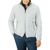 A person wearing a light gray, slim-fit zip-up jacket and dark blue pants is standing with hands in pockets. The Herno Olive Green White Reversible Nylon Blouson, made of water-resistant nylon, features black zipper accents and a slightly raised collar.