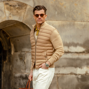 A man in a stylish Herno light camel wool silk nylon padded jacket and sunglasses stands confidently in front of an old stone archway, holding a red bag.