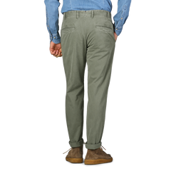 A person dressed in a blue shirt and Hiltl's Green Washed Cotton Gabardine Slim Chinos, seen from behind with hands tucked in pockets against a plain backdrop.