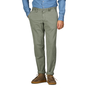 A person wearing a blue shirt, Green Washed Cotton Gabardine Slim Chinos by Hiltl, and brown shoes stands with hands in pockets.