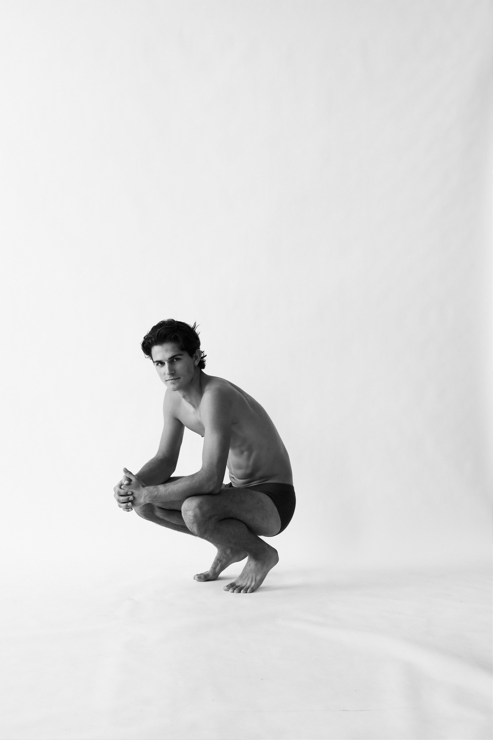 A person is squatting on a plain background, wearing dark Rio Pima Cotton Lyocell Briefs from The White Briefs, crafted from an ultra-soft blend of lyocell and Pima cotton, looking towards the camera. The image is in black and white.