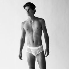 A man poses against a simple backdrop, displaying the Platan Organic Cotton Mesh Briefs from The White Briefs in a standard fit. The white mesh fabric enhances the monochrome photograph as he elegantly gazes to the side.