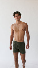 A person stands against a plain background, wearing The White Briefs' Henner Cotton Lyocell Boxer Shorts in olive green, featuring an elastic waistband and crafted from a lyocell-cotton blend.