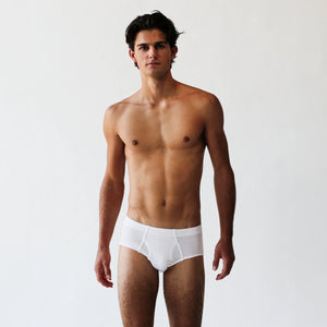 A man is standing against a simple backdrop, dressed in the Rio Pima Cotton Lyocell Briefs by The White Briefs, made from a luxurious blend of soft lyocell and Pima cotton.