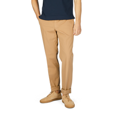 A man wearing Incotex Beige Cotton Stretch Regular Chinos, beige sneakers, and a navy blue shirt stands with hands in pockets, exuding casual elegance.