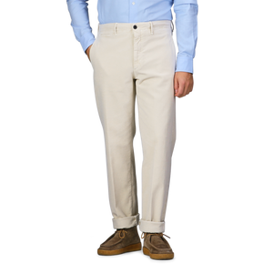 A person wearing Incotex Cream Beige Cotton Moleskin Slacks Chinos, a light blue shirt, and brown shoes stands with one hand in their pocket.