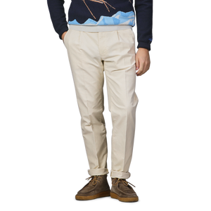 A person wearing a dark sweater, Cream White Cotton Twill Pleated Chinos by Incotex, and brown shoes stands against a gray background.