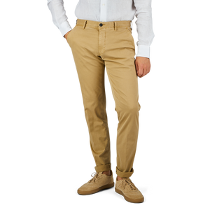A person stands with hands in pockets, wearing Incotex Dark Beige Cotton Stretch Slacks Chinos, a white long-sleeved shirt, and light brown shoes. The background is plain and white.