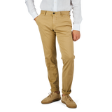 A person stands with hands in pockets, wearing Incotex Dark Beige Cotton Stretch Slacks Chinos, a white long-sleeved shirt, and light brown shoes. The background is plain and white.