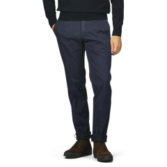 A person is wearing Incotex slim fit dark blue cotton stretch Model 30 chinos and brown shoes, with hands in pockets, against a plain background.