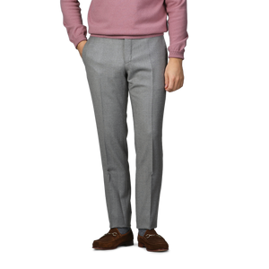 A person wearing a pink sweater, Light Grey Wool Flannel Slim Trousers by Incotex, and brown leather shoes stands with hands in pockets against a plain background.