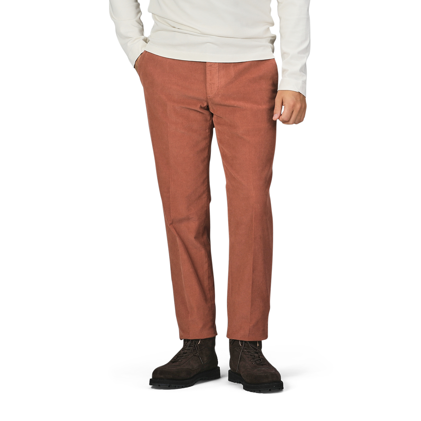 A person wearing a white long-sleeve shirt, Incotex Muted Orange Cotton Corduroy High Comfort Chinos, and dark brown boots stands with their hands in their pockets against a plain background.