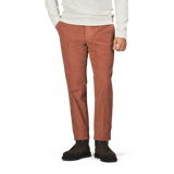 A person wearing a white long-sleeve shirt, Incotex Muted Orange Cotton Corduroy High Comfort Chinos, and dark brown boots stands with their hands in their pockets against a plain background.