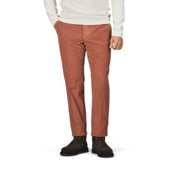 A person wearing a white long-sleeve shirt, Incotex Muted Orange Cotton Corduroy High Comfort Chinos, and dark brown boots stands with their hands in their pockets against a plain background.