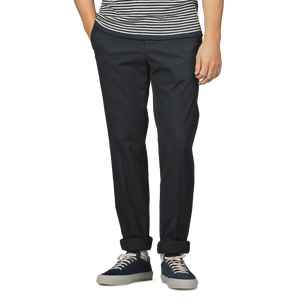 Someone wearing a striped shirt, Incotex Navy Blue Cotton Royal Batavia Regular Chinos, and dark sneakers stands with one hand in their pocket.