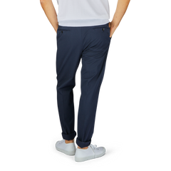 A person standing with their back to the camera, wearing a white shirt, navy blue Navy Blue Cotton Stretch Regular Chinos from Incotex, and white sneakers. Hands are placed in the pants' pockets.