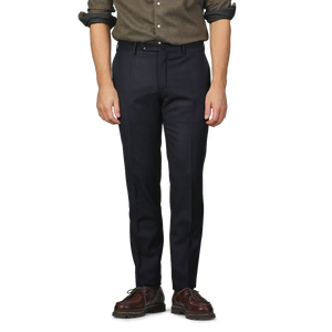 A man dressed in Incotex Navy Blue Wool Flannel Slim Trousers, a brown shirt, and brown shoes stands with his arms by his sides against a plain background. The slim fit of the trousers highlights his relaxed yet refined stance.