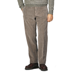 A person in Incotex's Stone Grey Cotton Corduroy Regular Fit Trousers and brown shoes, paired with a gray sweater, stands with hands in pockets against a plain background.