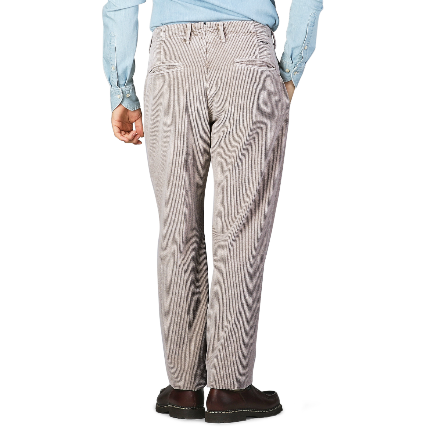 A person wearing taupe beige cotton corduroy regular fit trousers from Incotex, a light blue shirt, and brown shoes is standing with a casual stance, facing away from the camera.