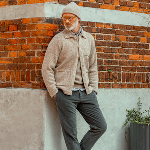 A man wearing an Inis Meain Beige Melange Wool Cashmere Carpenter Jacket is casually leaning against a brick wall.