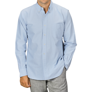 A man wearing a Light Blue Washed Cotton Oxford BD Shirt from Kamakura Shirts and gray trousers. The pure cotton oxford shirt has a chest pocket, and the man has his hands in his pockets.