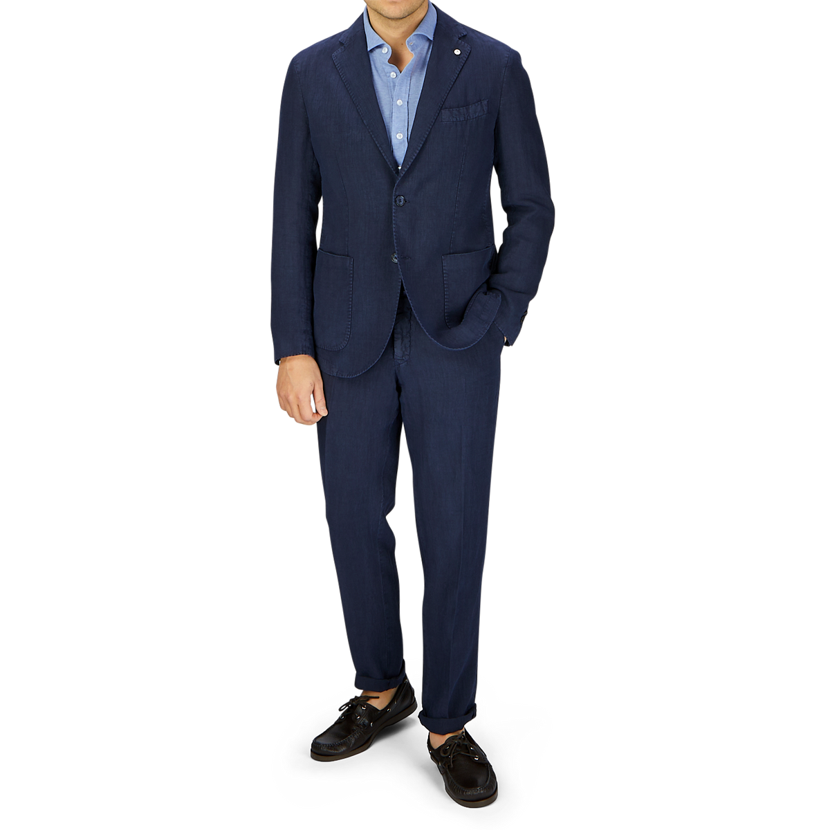 A man stands wearing a L.B.M. 1911 navy blue washed linen suit, blue shirt, and brown shoes, with his left hand in his pocket, against a grey background.