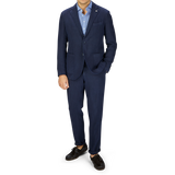 A man stands wearing a L.B.M. 1911 navy blue washed linen suit, blue shirt, and brown shoes, with his left hand in his pocket, against a grey background.