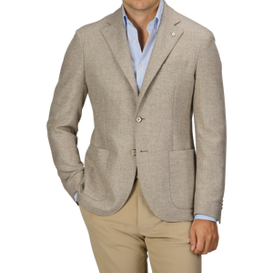 A man wearing an Oatmel Beige Shetland Wool Cashmere Blazer by L.B.M. 1911, paired with a light blue dress shirt and beige pants, stands with his hands in his pockets. The slim fit, seasonal blazer features two buttons and patch pockets.