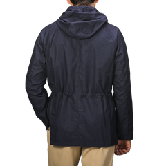 An individual is seen from behind against a plain background, wearing the L'Impermeabile Navy Blue Waxed Cotton Bob Short Parka and beige pants.