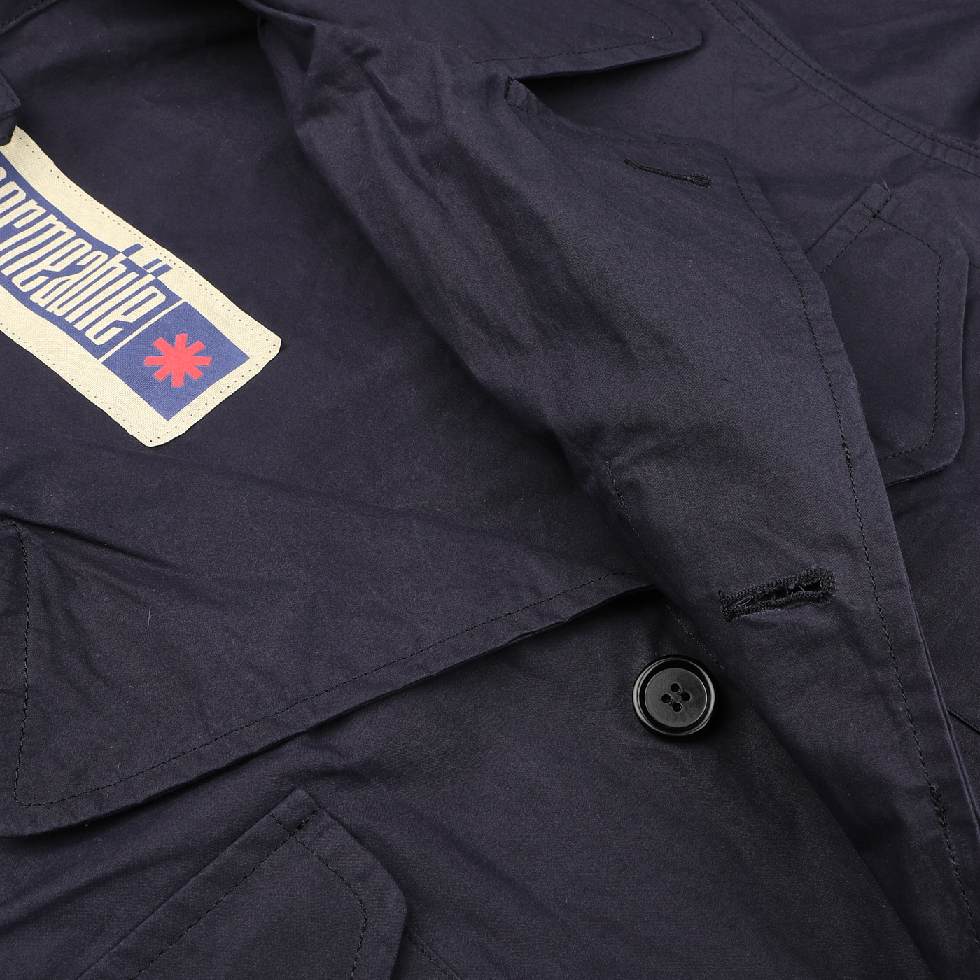 Close-up of a Navy Blue Waxed Cotton Smith SFO Jacket from L'Impermeabile, featuring two buttons and a patch with text and a red star-like symbol, designed to be water-repellant.