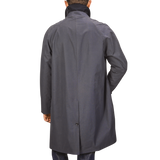 A person is standing with their back to the camera, wearing a knee-length Washed Blue Waxed Cotton Kamikaze Coat with a high collar and long sleeves from L'Impermeabile, made from wind and water-resistant waxed cotton.