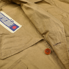 A close-up of the Khaki Beige Waxed Cotton Smith SFO Jacket by L'Impermeabile showcases Italian craftsmanship with a wooden button and stitched blue-red label. It combines elegance and practicality, featuring a subtle water-repellent finish.