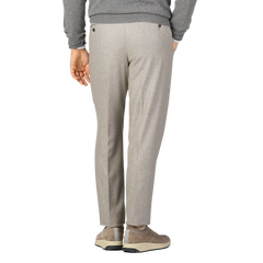 Person wearing oatmeal beige Luigi Bianchi wool flannel flat front trousers, a gray sweater, and beige sneakers stands with one hand in their pocket, facing away from the camera.